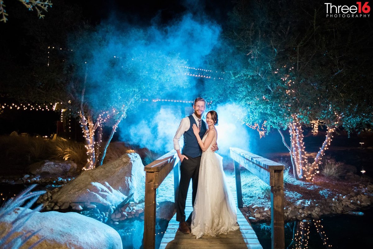 Bride and Groom pose for a night photo while standing on a small bridge over a creek and a blue mist in the background