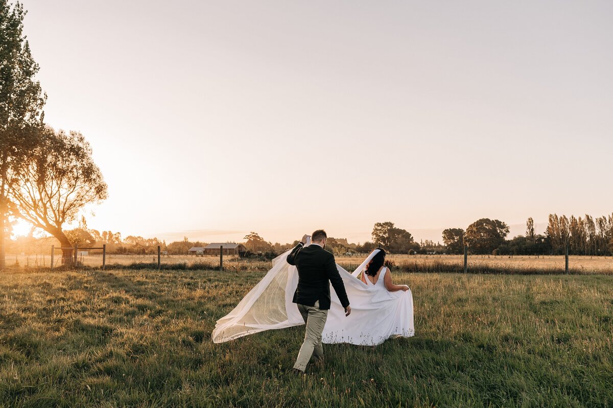 bride and groom walking across grass at sunset on farm in ohoka christchurch groom holding bride's train