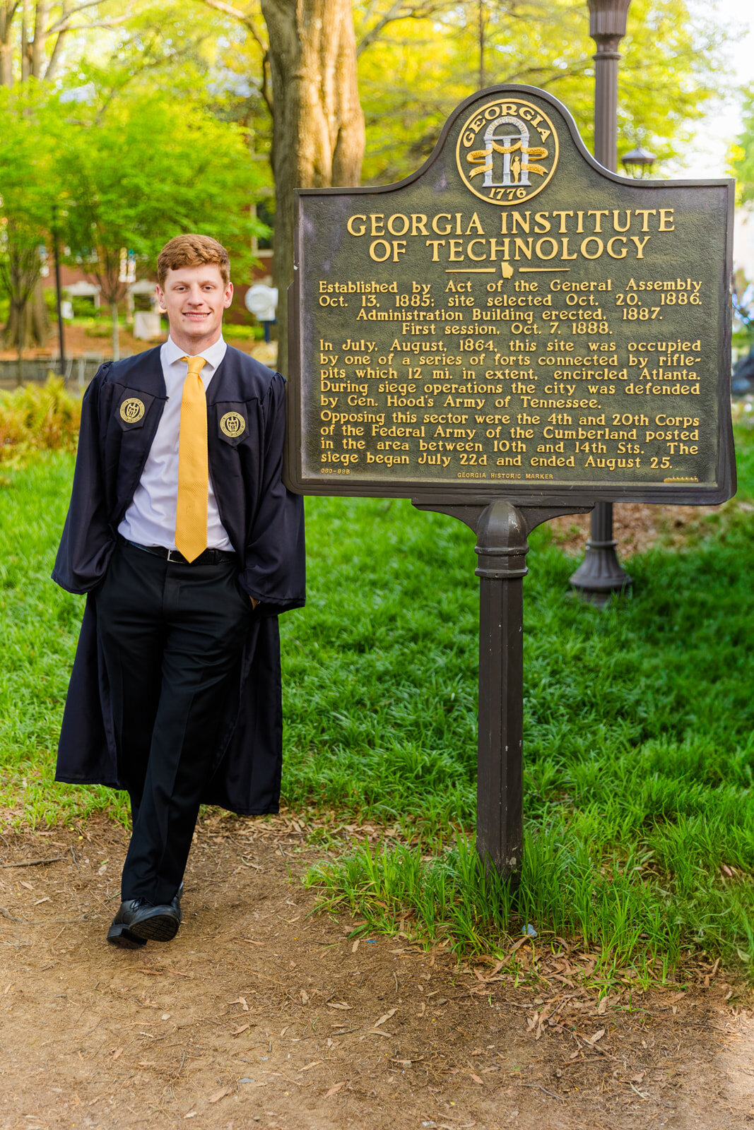 College graduate ankles crosses and hands in pockets wearing graduation gown leaning on the Georgia tech university history sign on campus during senior portraits with Atlanta photographer Laure Photography
