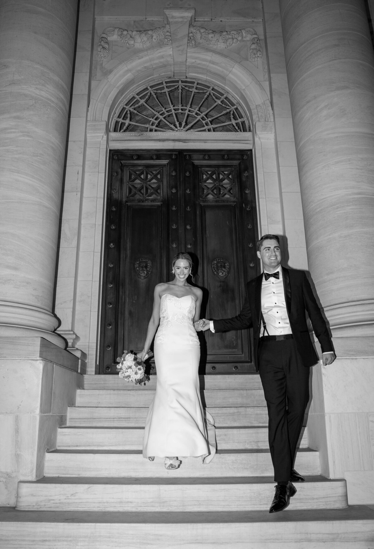 A bride and groom walk down steps in front of a grand building with large wooden doors. The bride wears a strapless gown and holds a bouquet, while the groom is in a tuxedo. The image is in black and white.