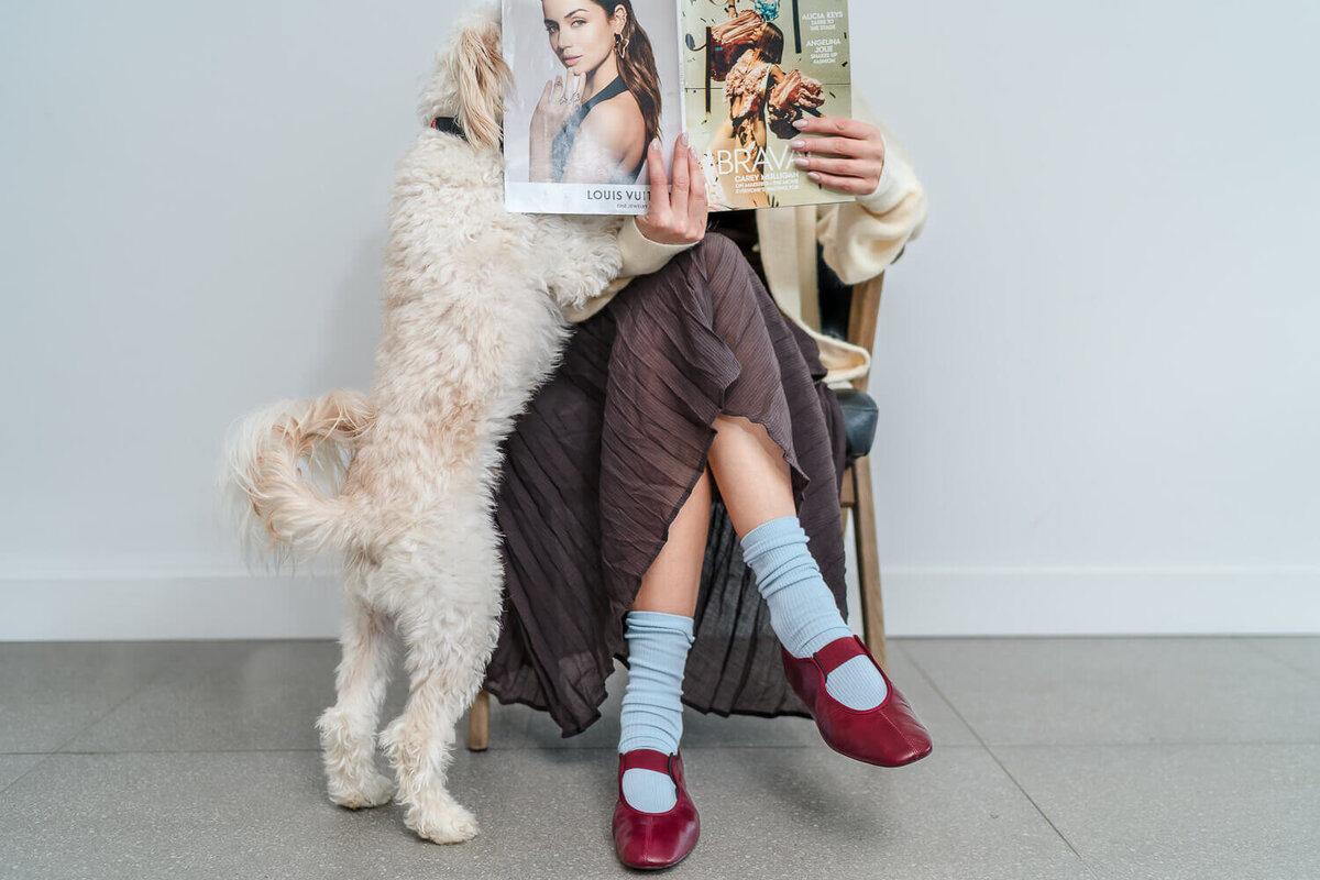 White fluffy dog jumping on seated woman reading magazine with red shoes and pleated skirt.