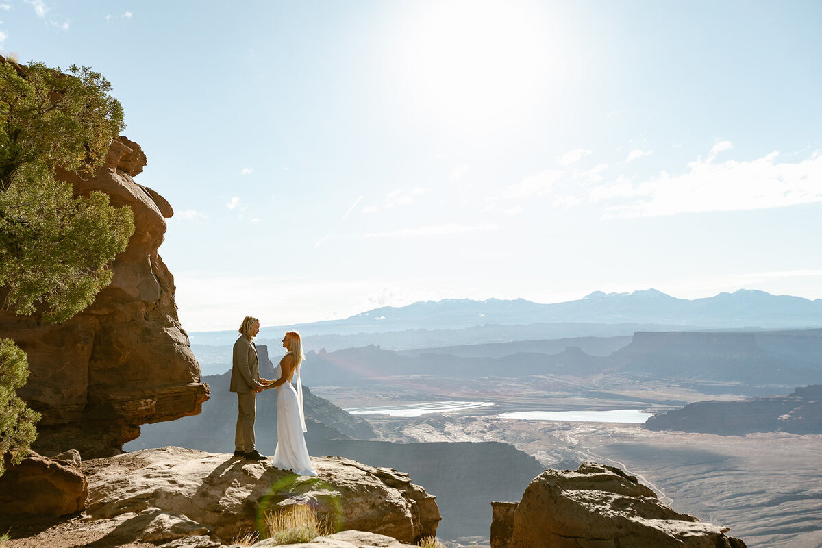 A couple standing holding hands  at Deadhorse State Park in Moab, on their elopement day as the sunrises. 