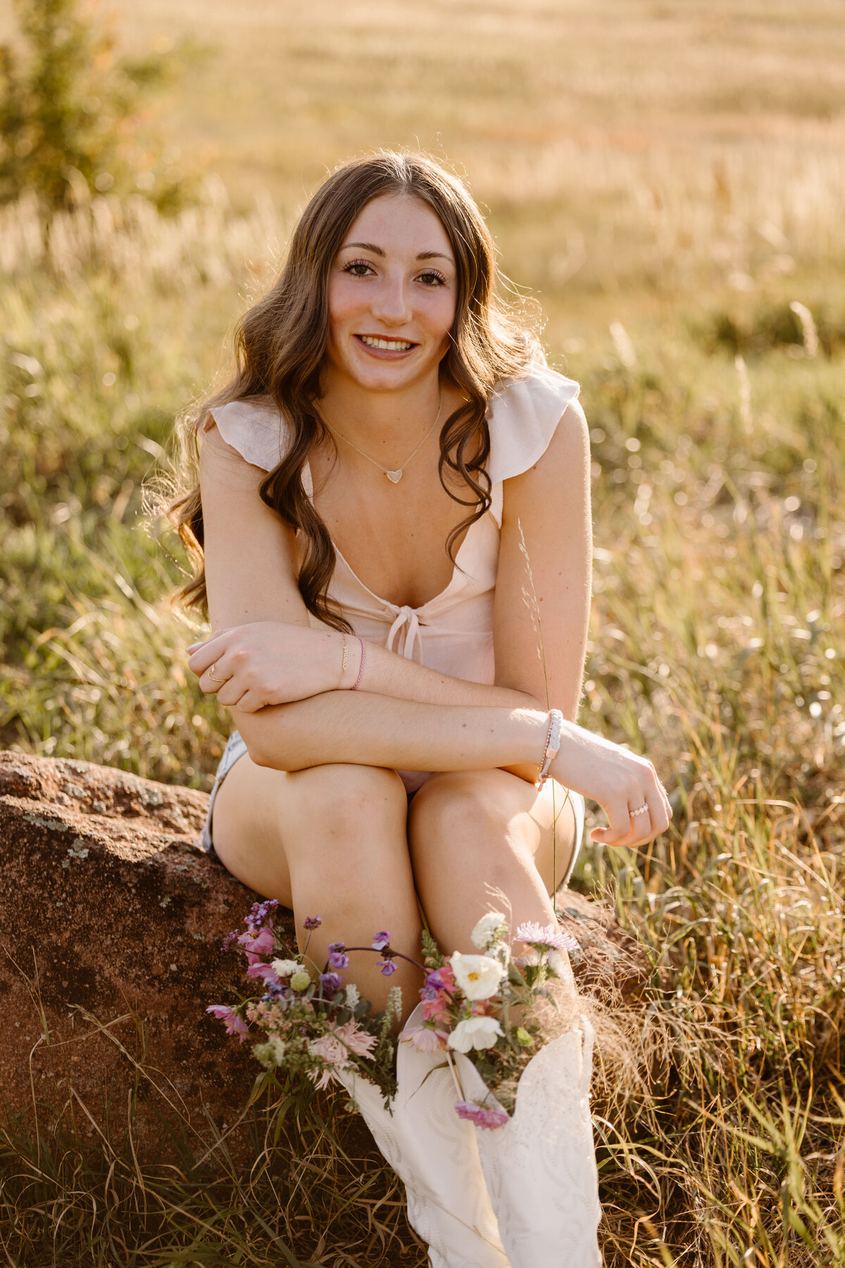 Teen girl sits on a rock during golden hour wearing white boots filled with wildflowers, smiling warmly in a field of tall grass during her senior photo session in Boulder, Colorado.