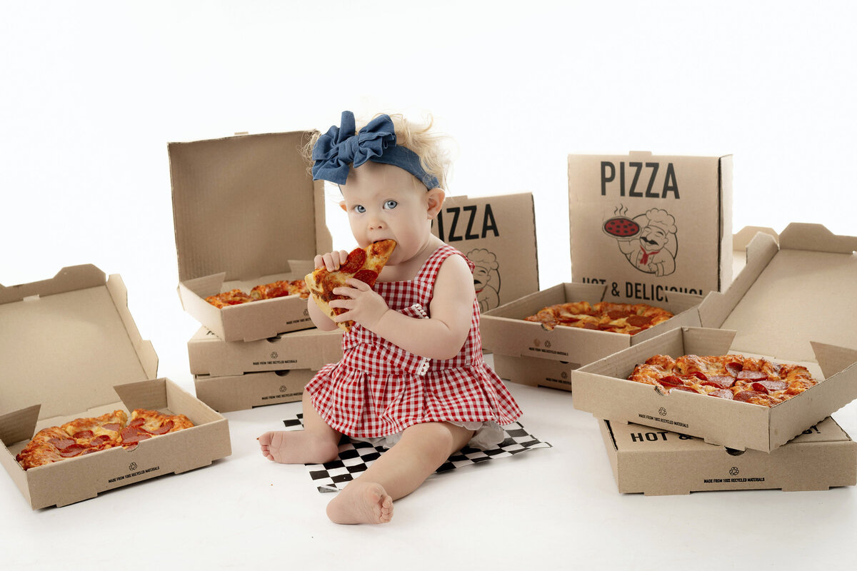 Toddler sitting among pizza boxes chewing on a slice.
