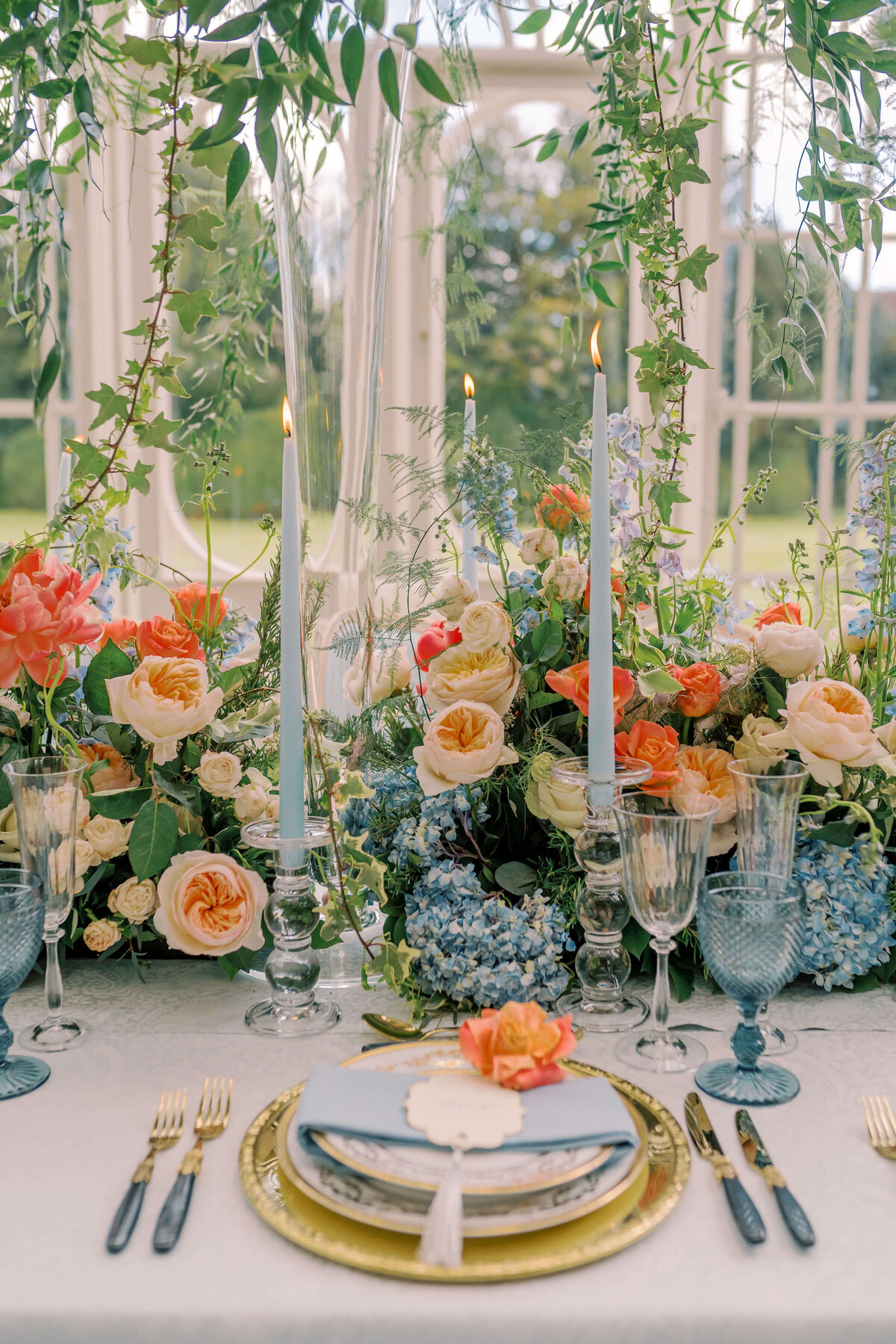 wedding dinner table with a blue and coral colour scheme, gold plates, and blue water glasses in avington park’s orangery overlooking the gardens