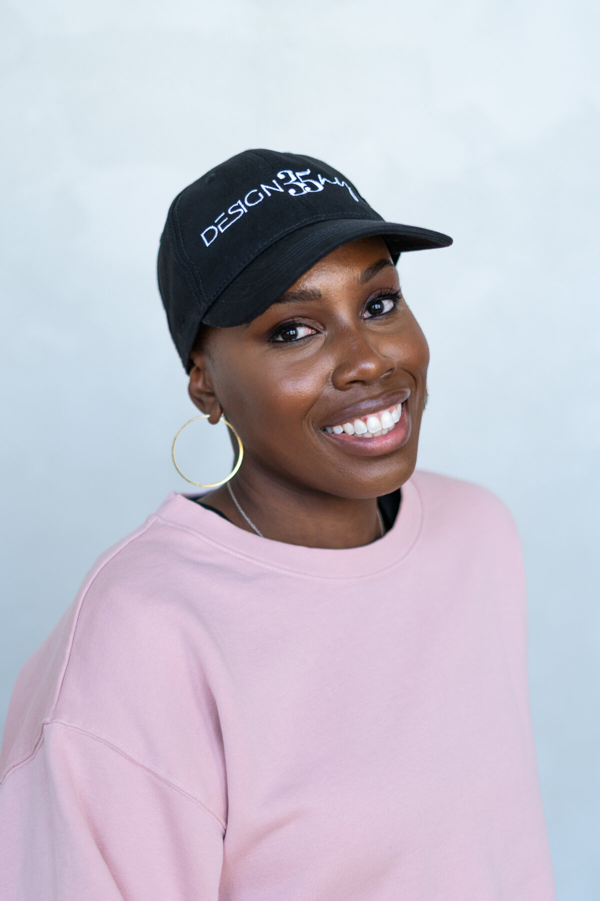 A clean headshot of the artist in her branded hat, set against a soft, dabbled painted backdrop. This portrait captures her warmth and professionalism in a polished, editorial style.