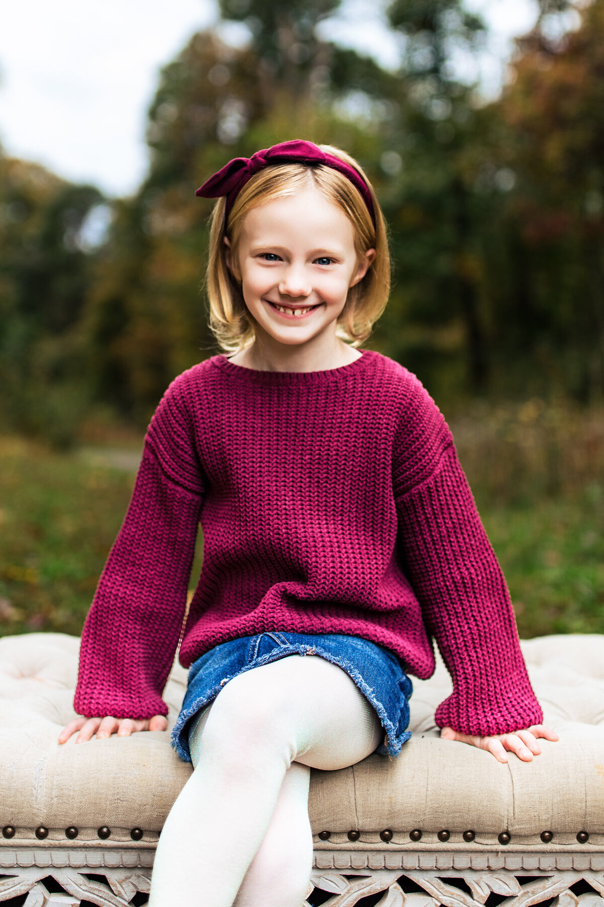 A blond girl with a bright Burgundy sweater and a bright Burgundy bow sits on an ottoman during a woodland photography shoot in the woods around Pittsburgh Pennsylvania