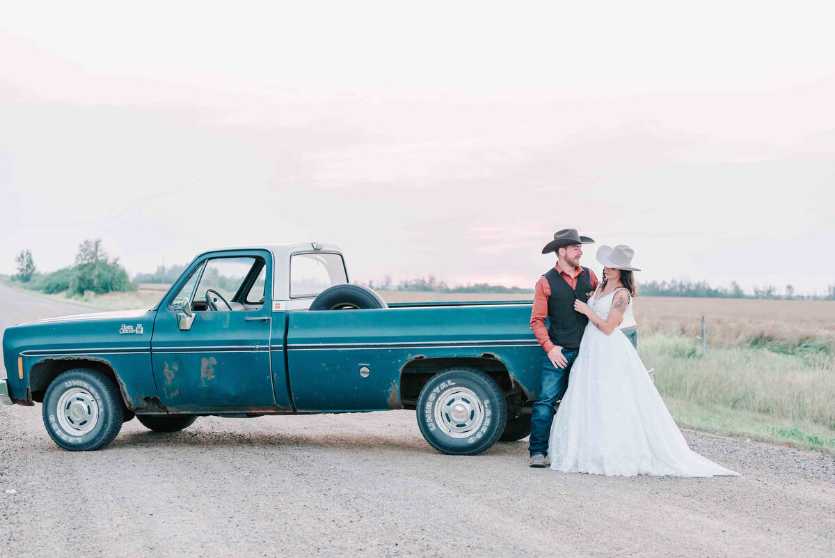 Groom leans against the tailgate on the green truck. Bride faces him. 
