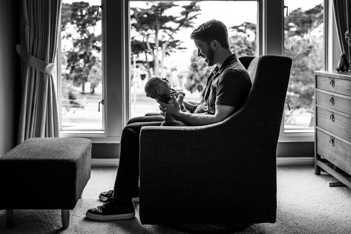 dad sitting in rocker in front of window holding baby and looking at him smiling