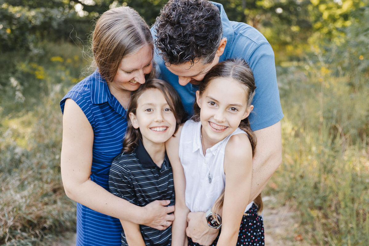 Family of four smiling and hugging outdoors with greenery in the background. Captured in High Park Toronto.