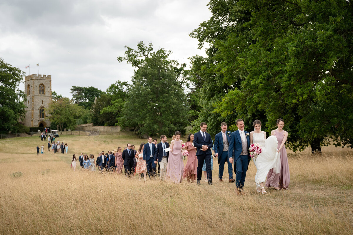 Just married couple leading their guests like the Pied Piper across a field with church in background