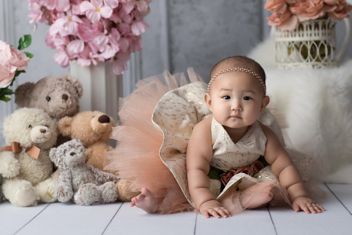 A baby girl dressed in a golden lace dress with a blush tulle skirt and a pearl headband poses gracefully among soft teddy bears and pastel floral arrangements. Captured by a Dallas baby photographer known for luxurious milestone photography.