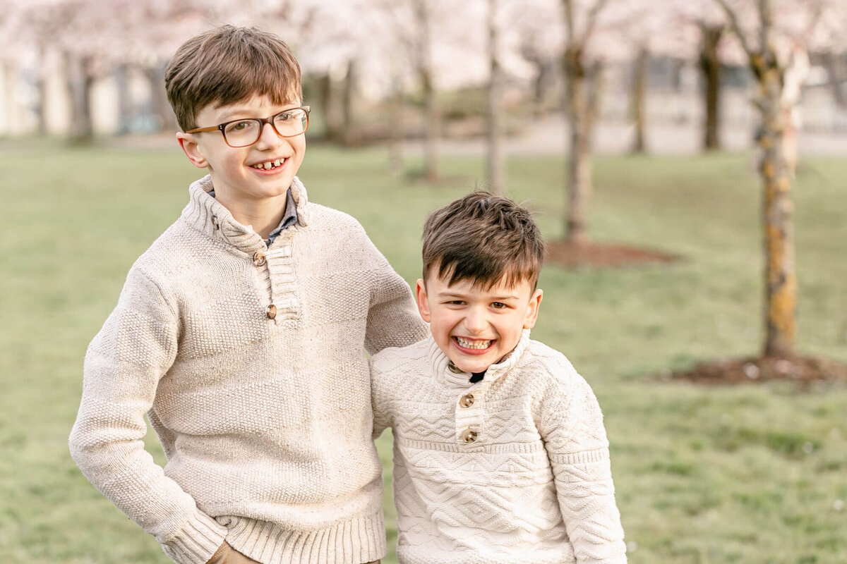 Two brothers in cream sweaters standing with their arms around each other and laughing towards the camera. They are standing in front of a row of cherry blossom tress. They are about 8 and 5 years old.