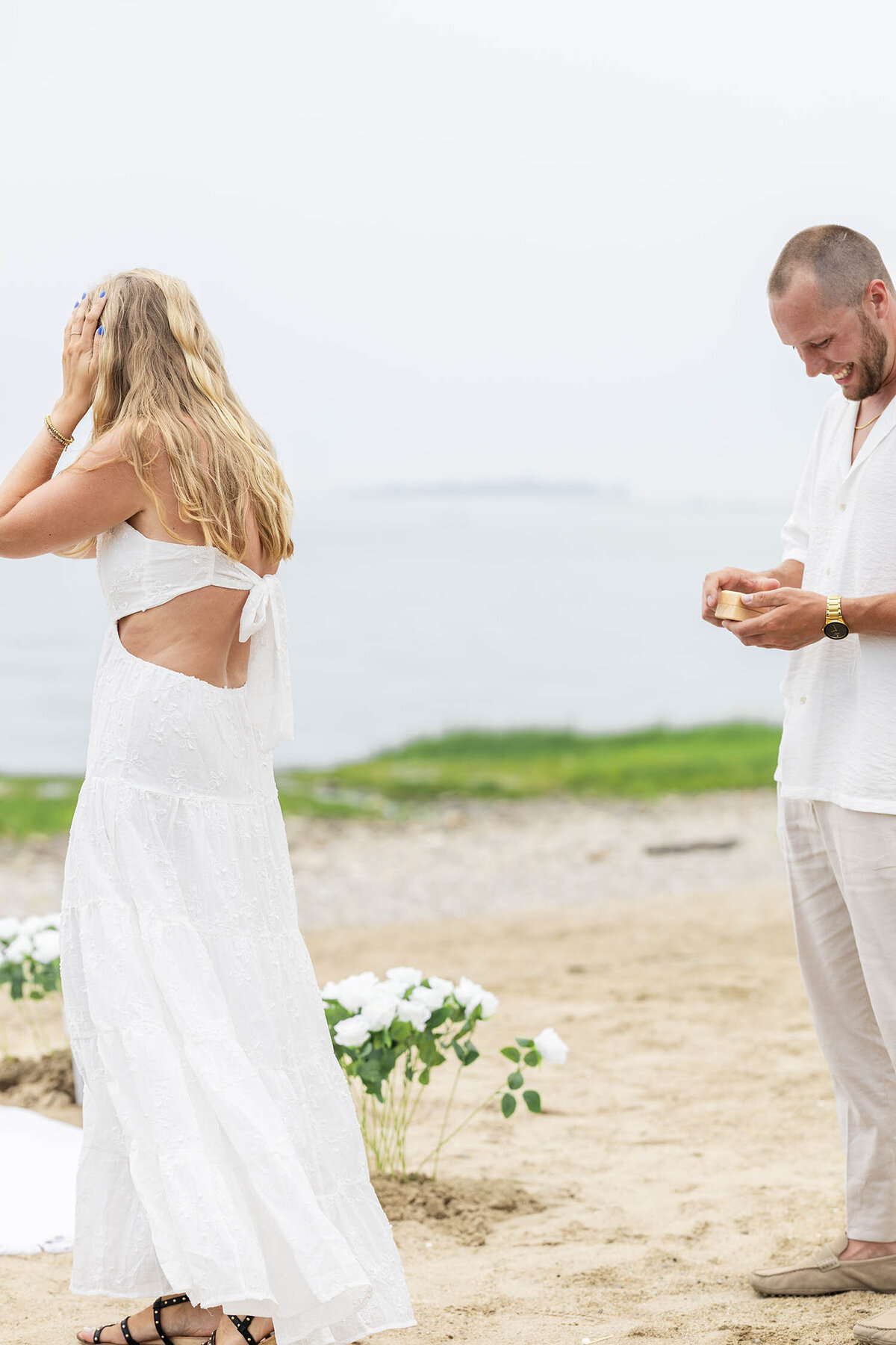 Woman wearing a white dress has her hands on her head as man wearing tan shirt and cream pants is smiling with a  ring box in his hand during a romantic Coastal Connecticut session. 