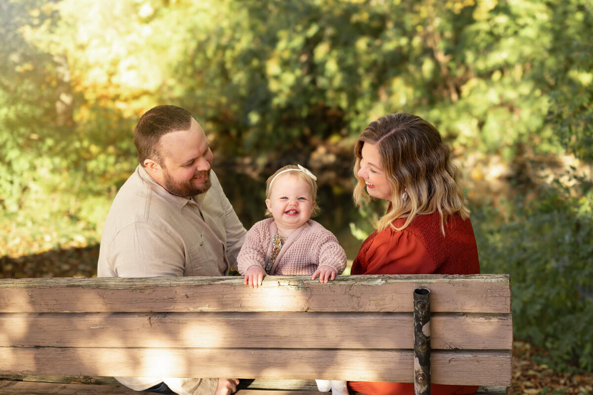 Parents sit on a bench with their toddler daughter in the middle of them in Michigan park