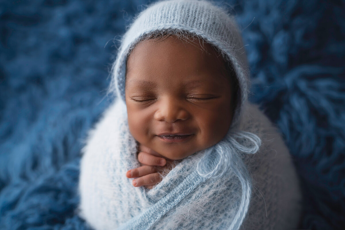 Sleeping baby boy is swaddled in light blue knit wrap and matching bonnet in penguin pose. Baby's hands are peek out from the swaddle. Baby is sleeping but with a smile on their face. Captured by Bryn Mwar Philadelphia and Main Line photographer Liza Nicole.