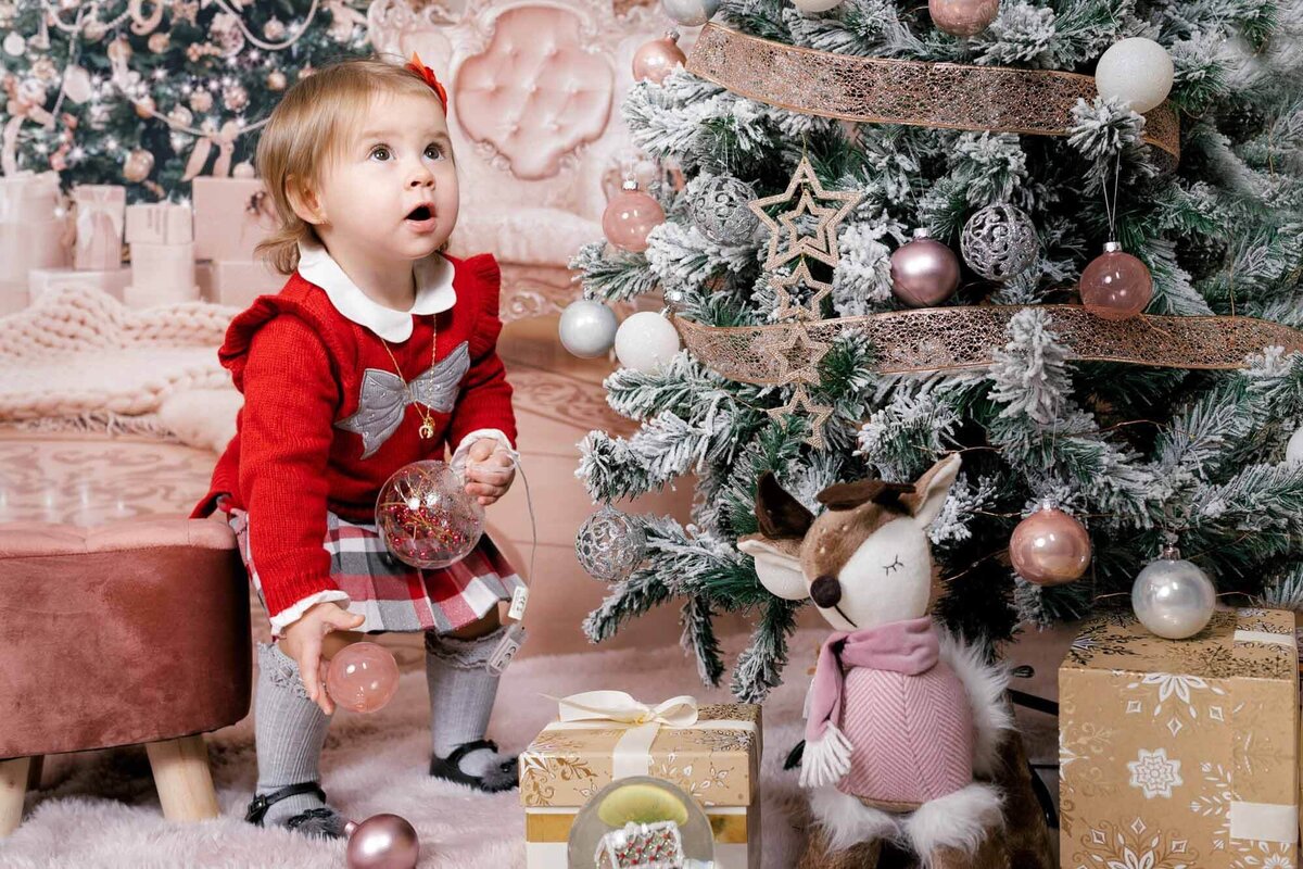 Toddler in red outfit playing with Christmas ornaments by the tree in Calini Weddings Studio - festive scene with wrapped gifts and soft golden lights.