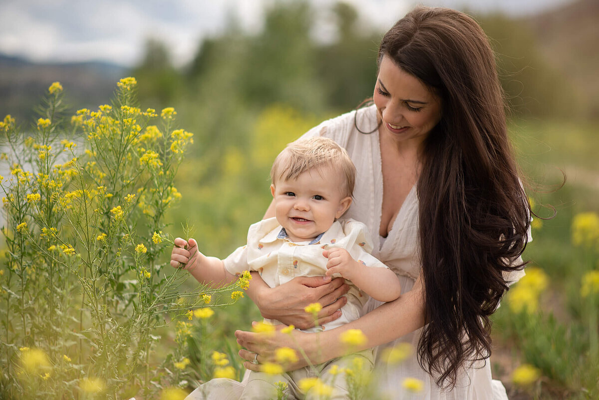 A mother and son sit in a field of yellow flowers. He sits on her lap and smiles and she looks down at him smiling. Her long brown hair flows by her side. Taken in Kamloops by Brittany Danielle Phtoography