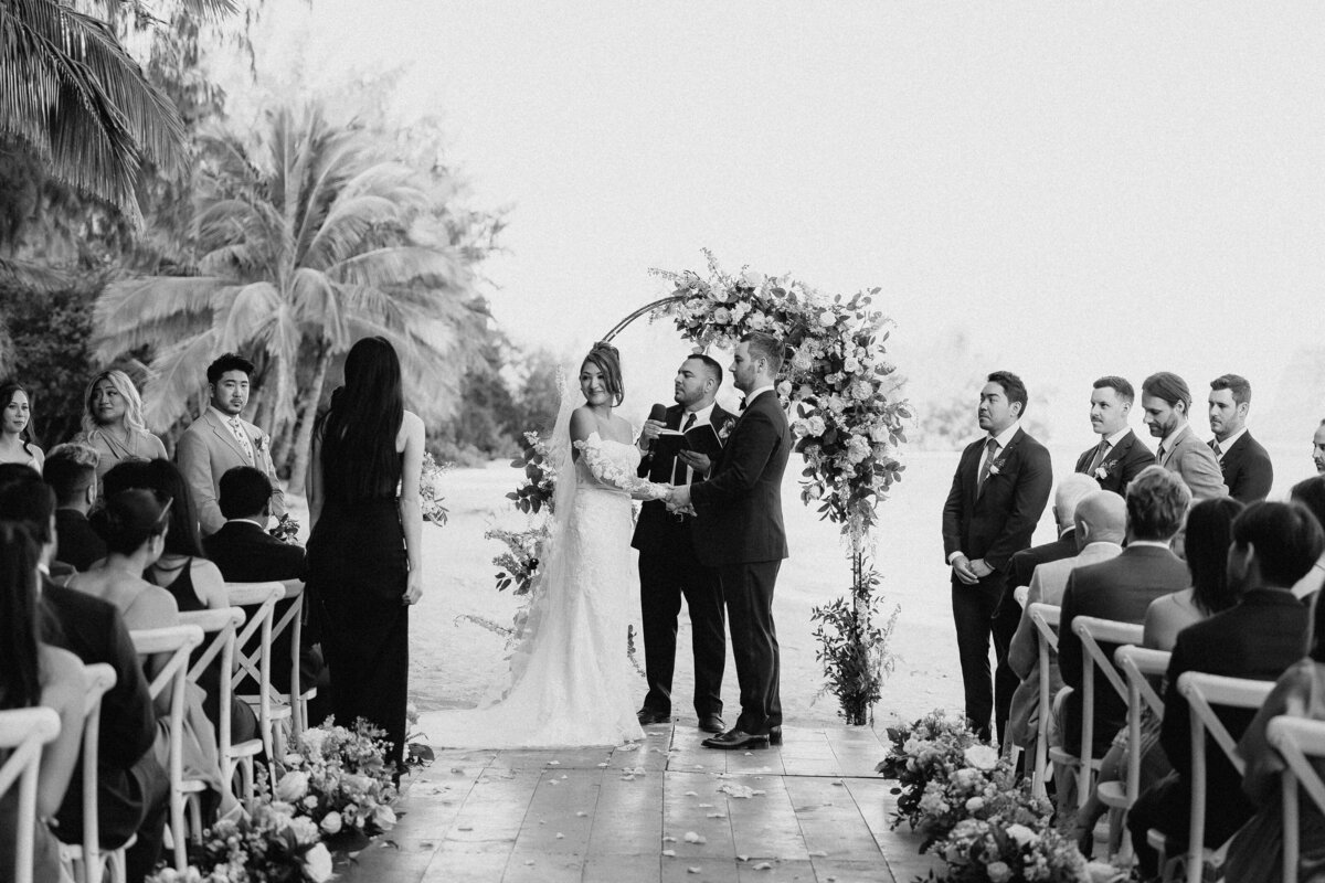 Bride and groom ceremony at Kualoa Ranch on the beach, holding hands with palm trees and ocean in the background in black and white