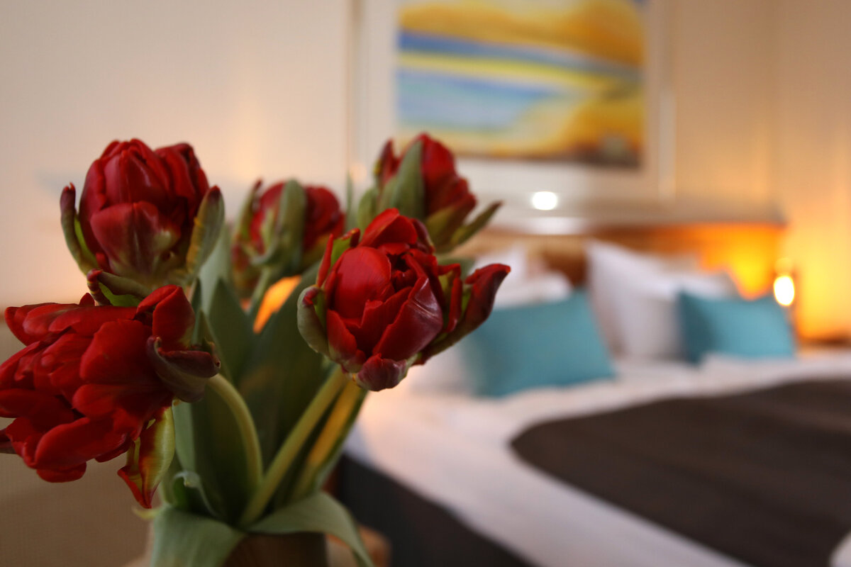 Ansicht eines Hotelzimmers, im Vordergrund sind Blumen und im Hintergrund ist das Bett zu sehen.