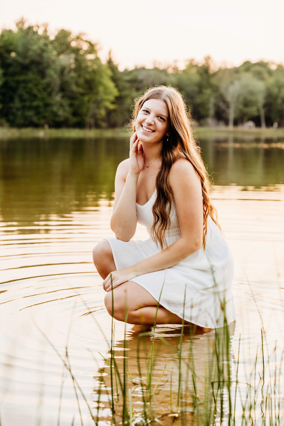 pretty teen girl in a white dress kneeling in the shallows and resting her chin on her hand as she enjoys her senior photography session with Ashley Kalbus.
