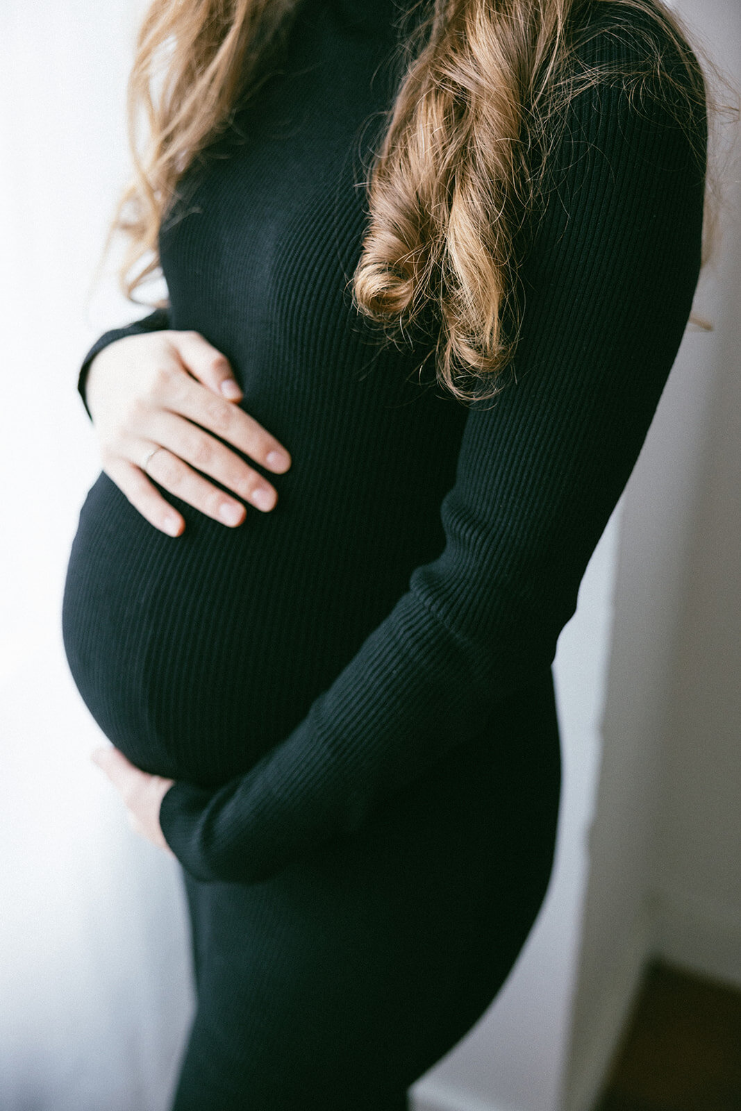 Pregnant woman rests her hand on her belly near a window in natural sunlight. 