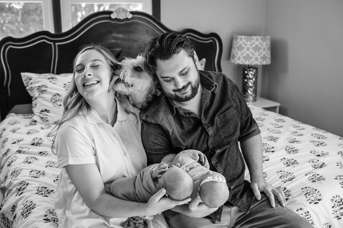 A black and white image of a mother and father sitting on a bed, holding their newborn twins.