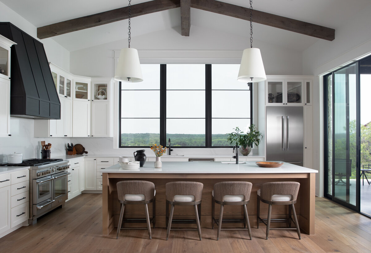 Hill Country Kitchen Remodel with matte black range, large wooden beams on vaulted ceilings White quartz countertops and brown wicker chairs. Stainless steel appliances.