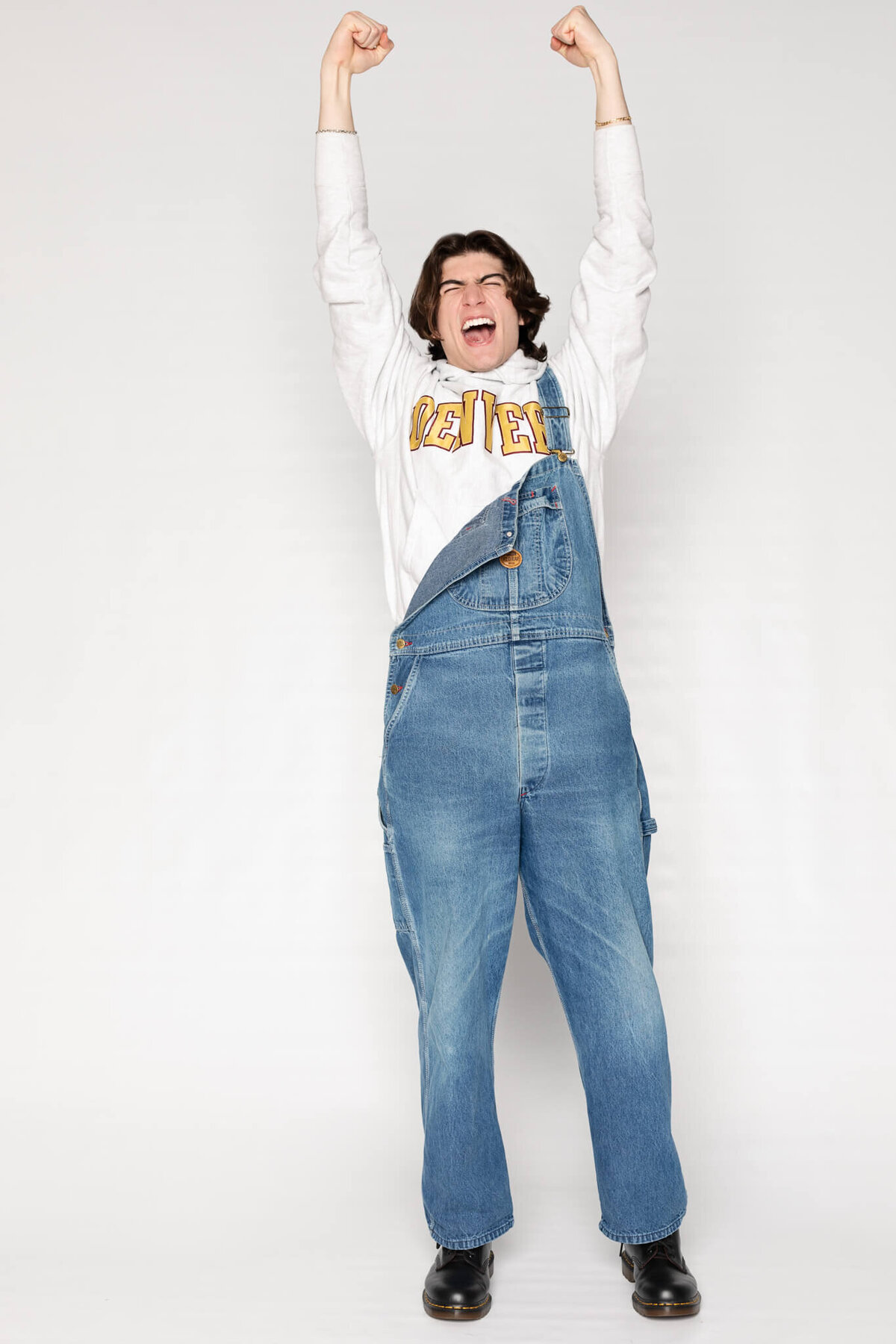 A person wearing denim overalls and a sweatshirt with CLEVELAND on it stands with arms raised and a joyful expression. The background is plain white.  .