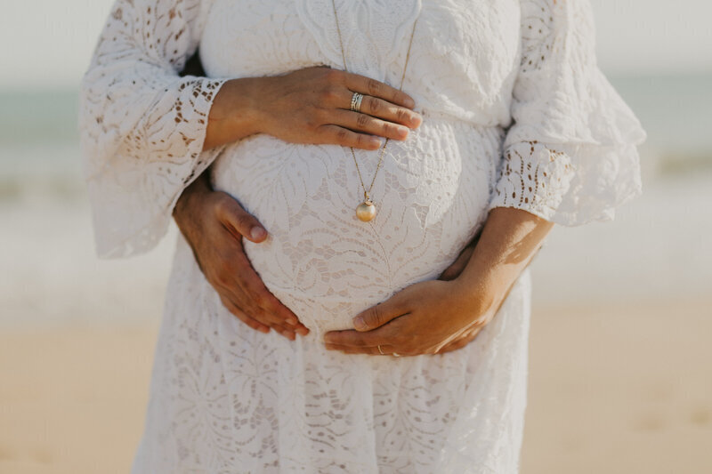 Quatre mains entourant le ventre d'une femme enceinte habillée avec une robe de dentelle blanche. Photo prise par Laura à la plage, photographe grossesse en Vendée.