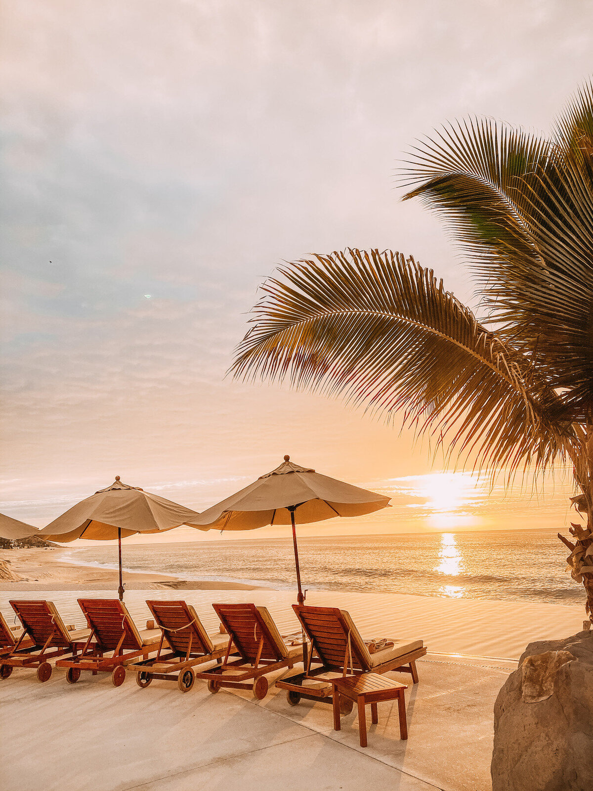 wooden lounge chairs at the beach at sunrise with umbrellas and a palm tree