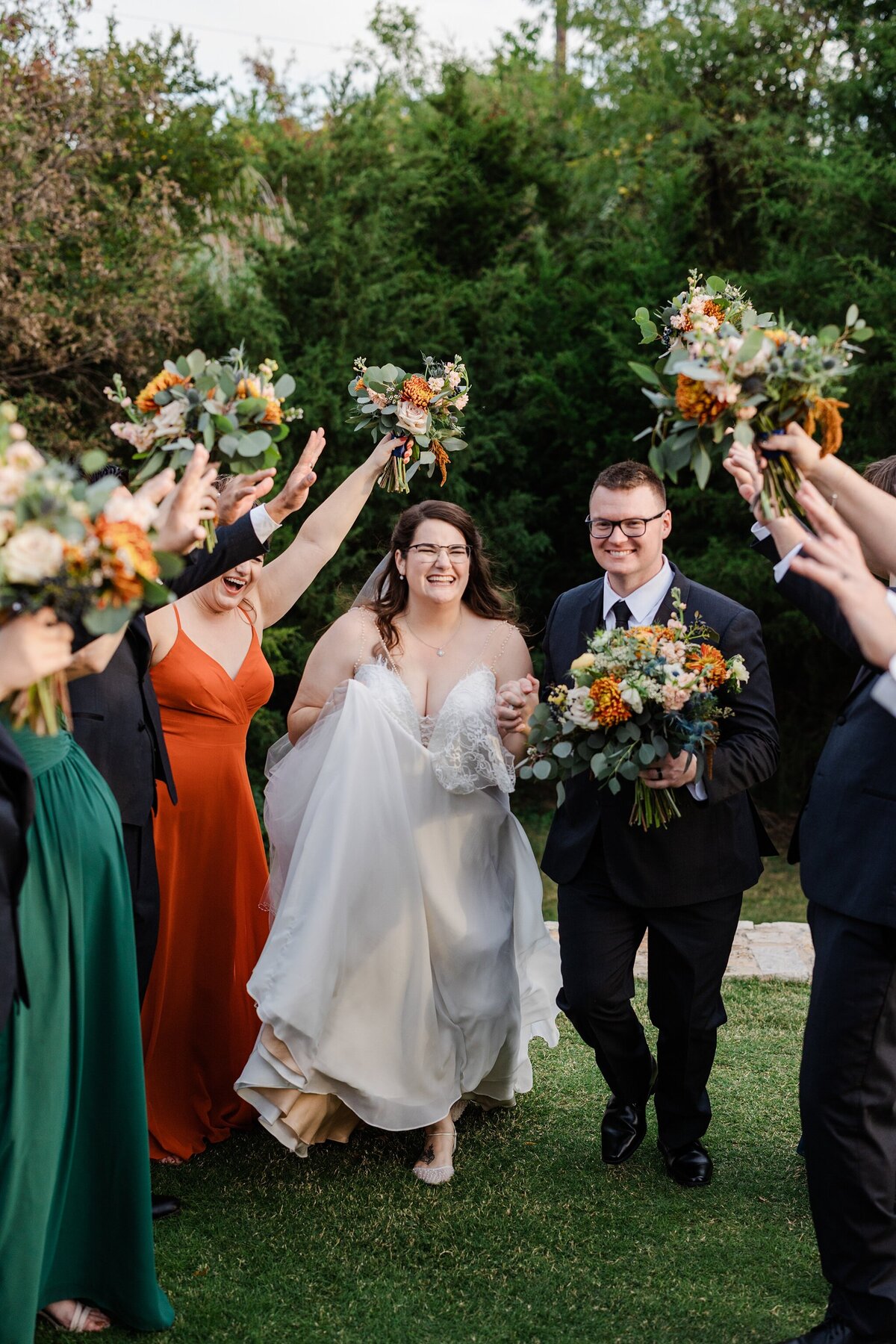 A portrait of a bride and groom walking through a "tunnel" made by their wedding party and their bouquets and arms after on their wedding day at The Laurel in Grapevine, Texas. The bride is on the left and is wearing a sleeveless, long, intricate, white dress. The groom is on the right and is wearing dark suit with a tie and is holding the bride's bouquet. Their wedding party can be seen on either side of them holding up their arms and bouquets in celebration.