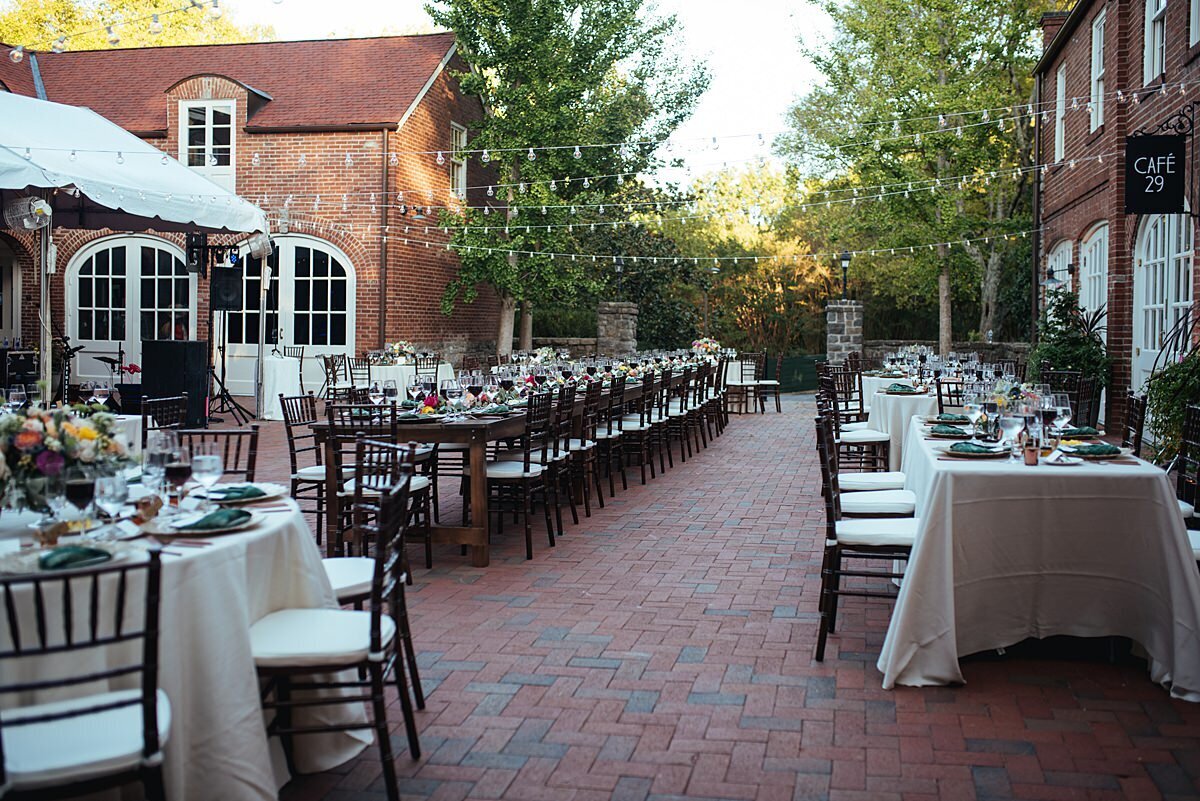 Wedding reception at the exposed brick Frist Learning Center courtyard at Cheekwood with long family style reception seating as well as round tables with ivory silk dupioni linen table cloths, large floral centerpieces, brown chiavari chairs and string lighting