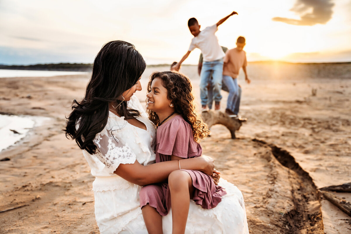 Family Photographer, a woman holds her daughter, her boys play behind them on the beach
