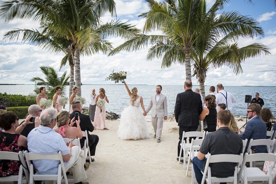 A bride and groom walk back up the aisle at the end of their tropical destination wedding, surrounded by palm trees. The bride throws her bouquet up in celebration.