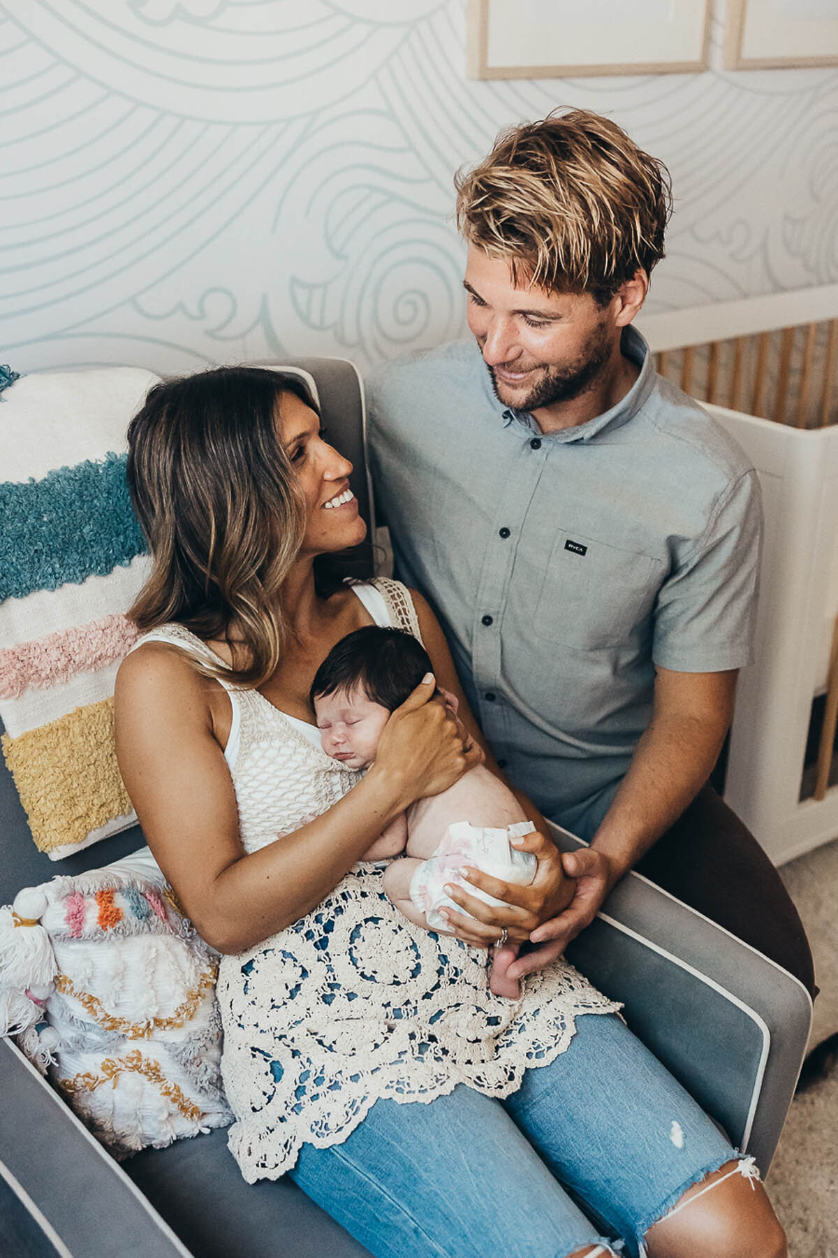 a mother holds her newborn baby while sitting in a chair and dad is next to them smiling at them