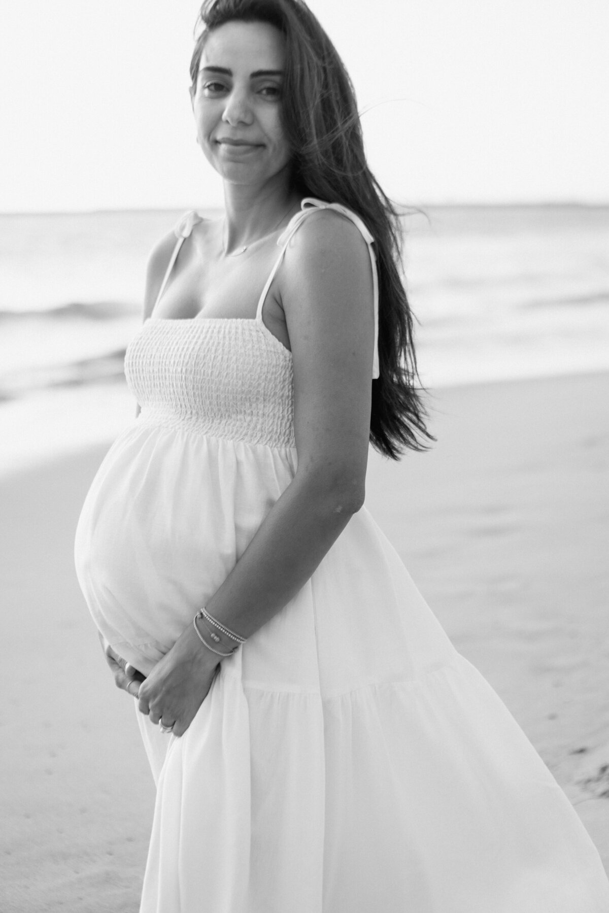 Pregnant mama cradles bump in black and white image on the beach during maternity session.