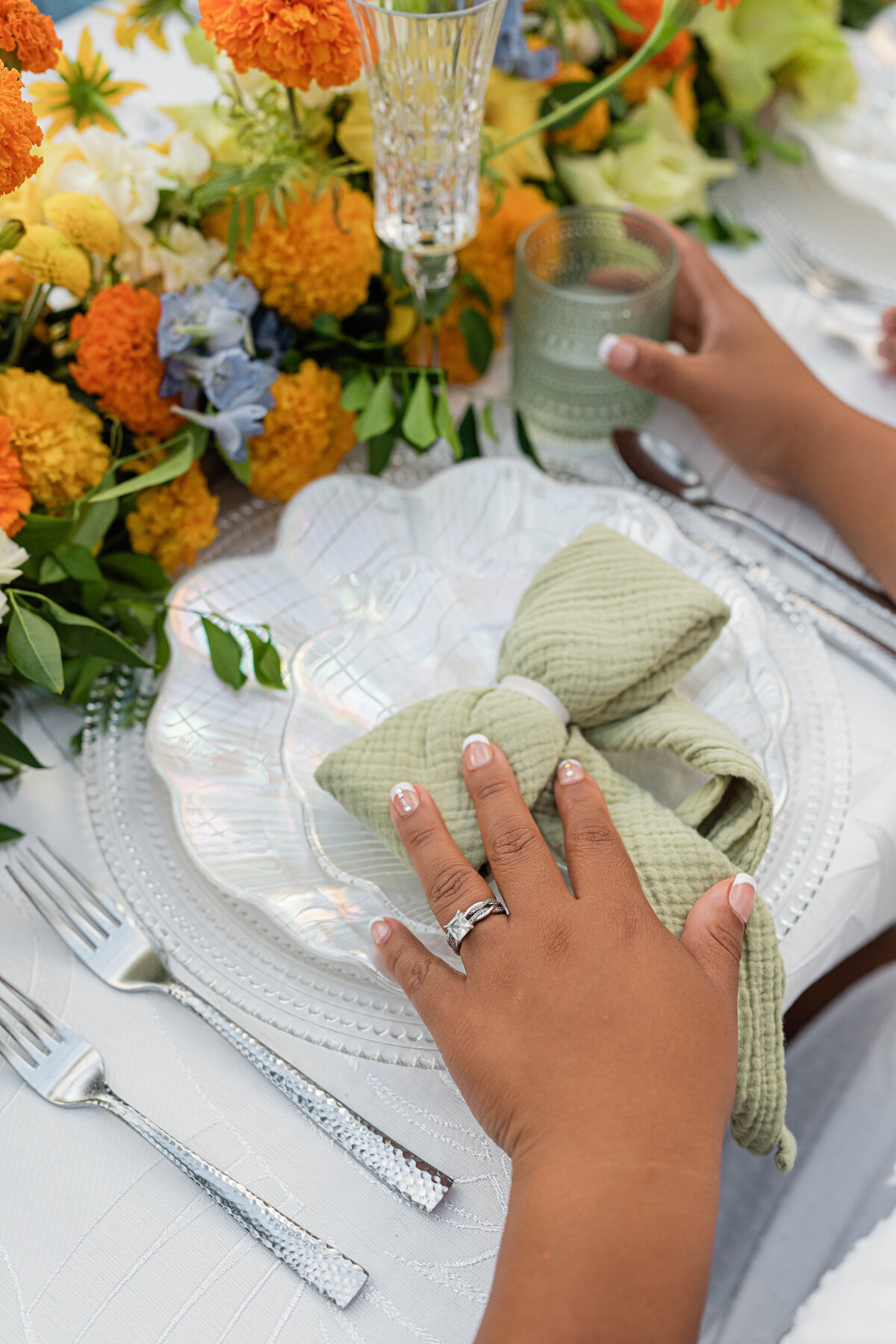 white on white palm leaf tablecloth adorned with a white, orange, blue and green floral table runner with hammered silver flatware there is a hand on an iridescent seashell plate placed on top of a clear beaded charger plate topped with a green gauze napkin tied in a bow