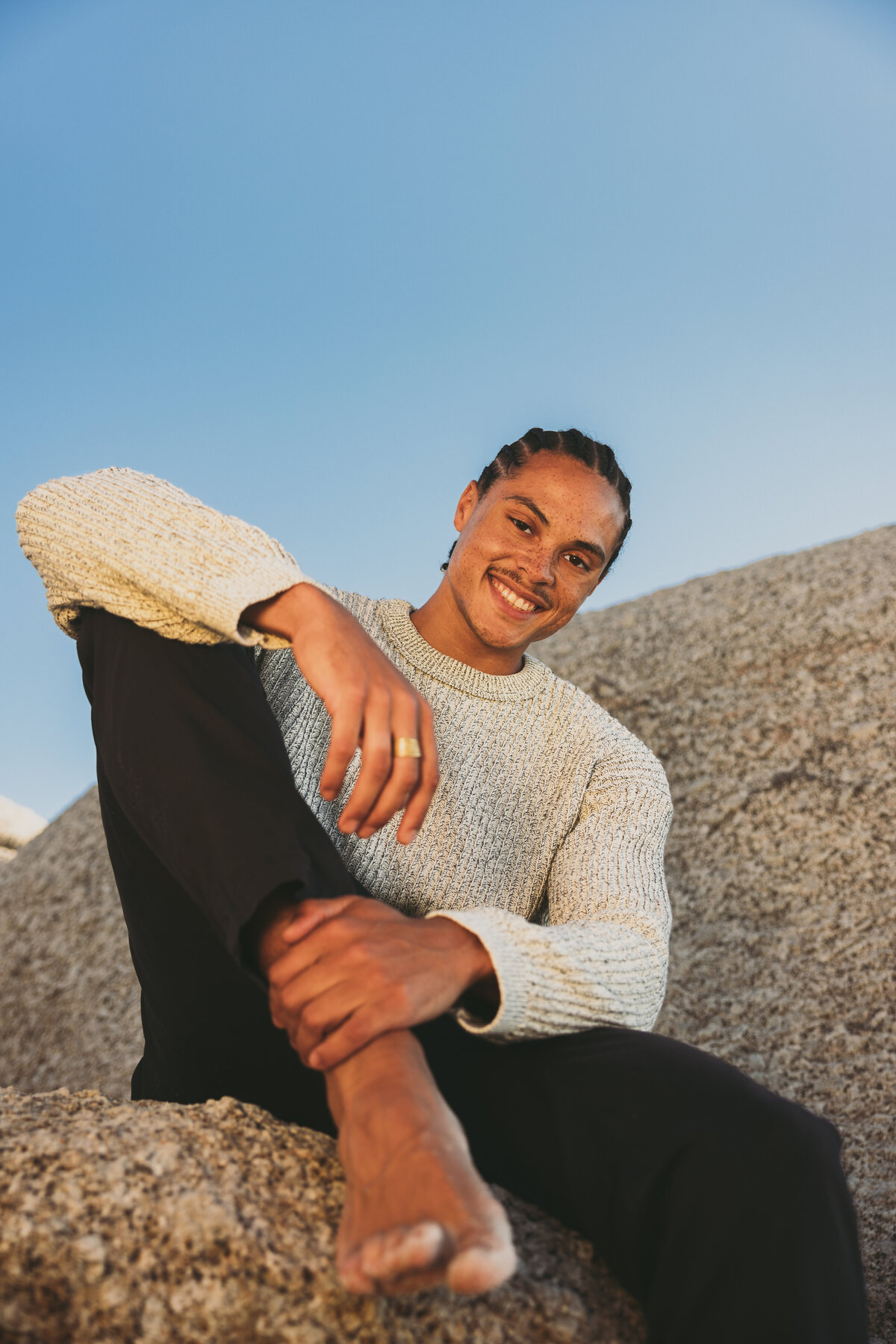 Portrait of Timothe sitting barefoot on a rock in Cape Town, smiling warmly while wearing a knitted sweater and black pants, framed by a clear blue sky.