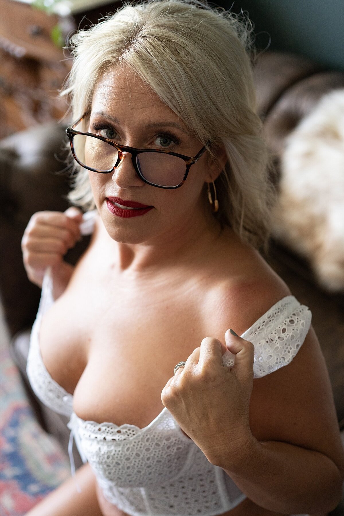 Mid-40's woman in a white peplum top with glasses posing seductively on the edge of a couch in Ferndale boudoir studio