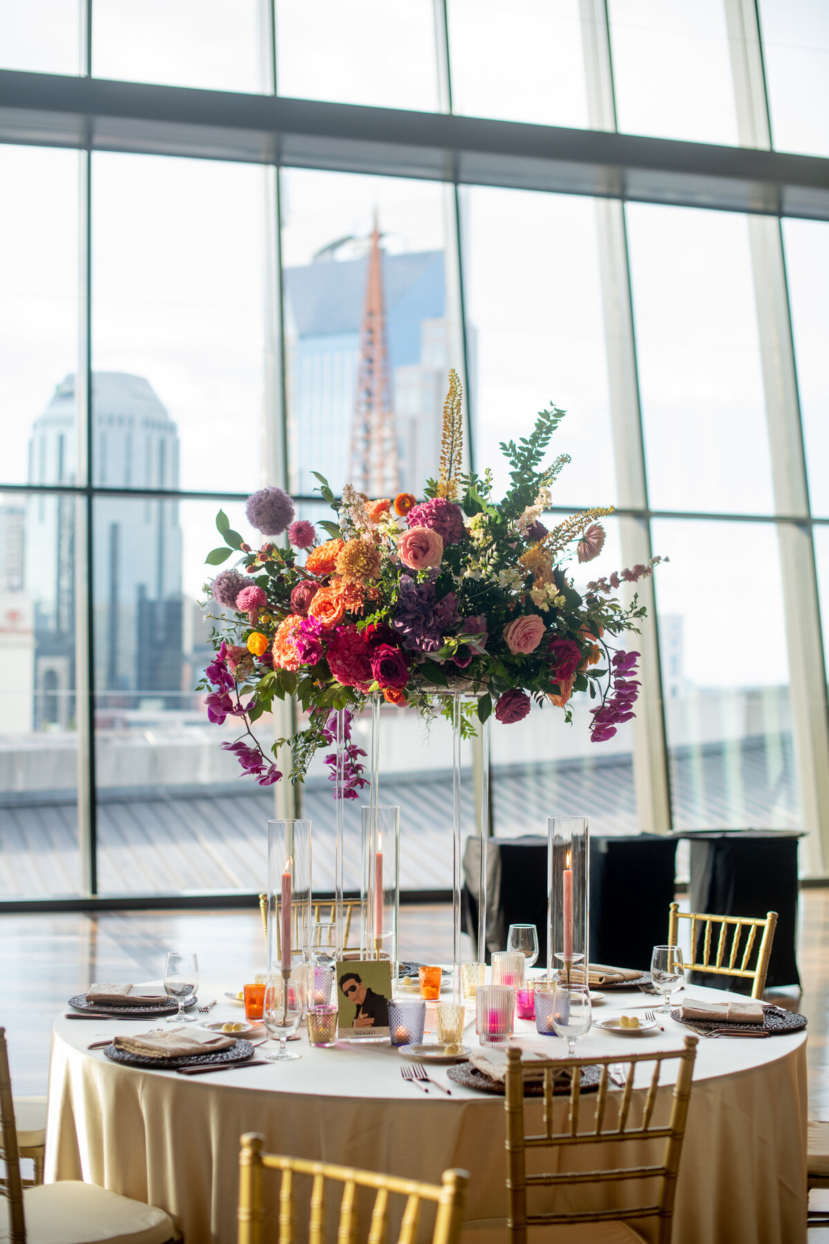 Lush elevated centerpieces in sunset hues of rosy pink, lavender, orange, and golden-yellow composed of phalaenopsis orchids, petal heavy roses, allium, delphinium, eremurus and natural greenery. Design by Rosemary and Finch in Nashville, TN.