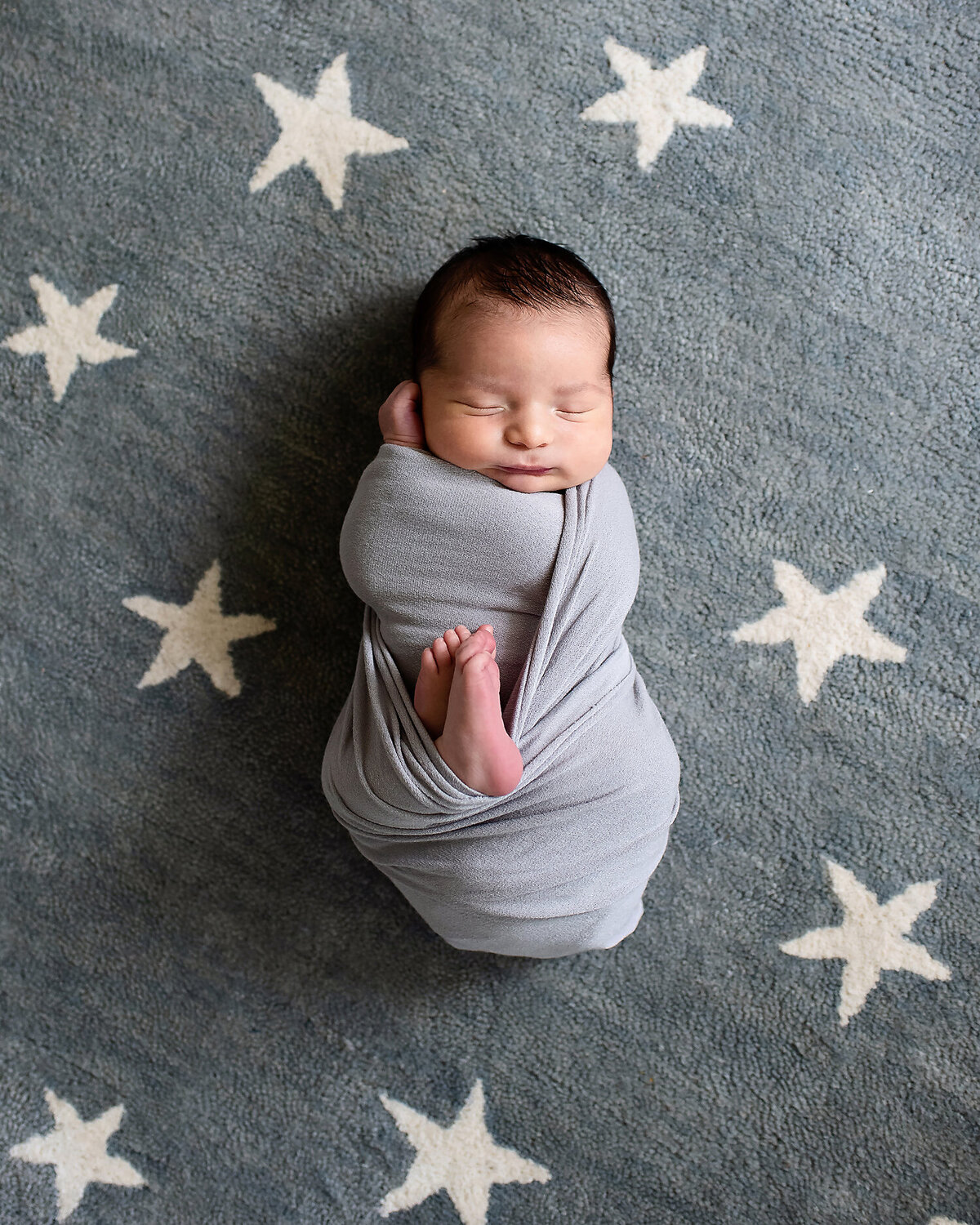 Newborn boy wrapped in a blue/gray swaddle laying on a rug with white stars in his newborn nursery.
