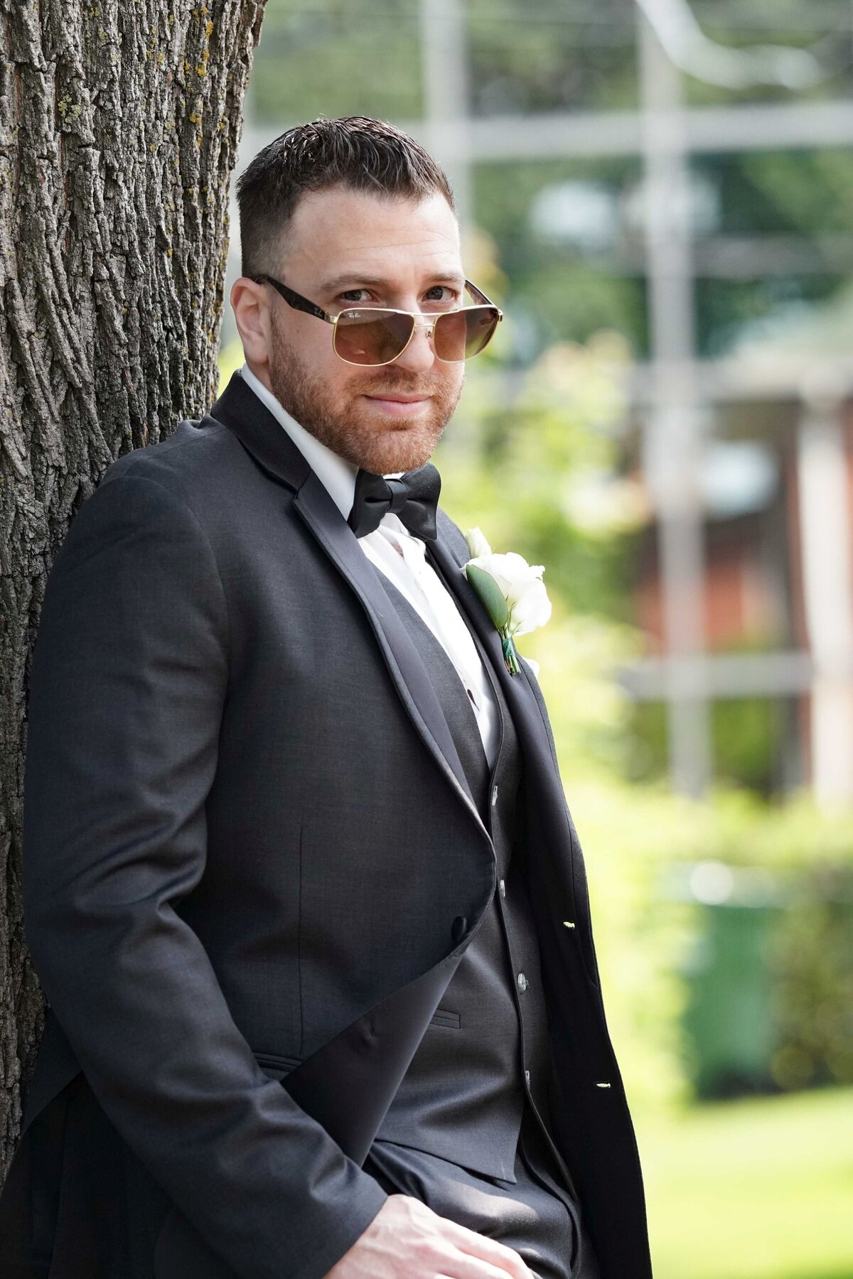 The groom leans casually against a tree, sporting sunglasses, which adds a relaxed and stylish flair to his look. This image captures a cool, confident moment, showcasing the groom's modern and laid-back attitude.