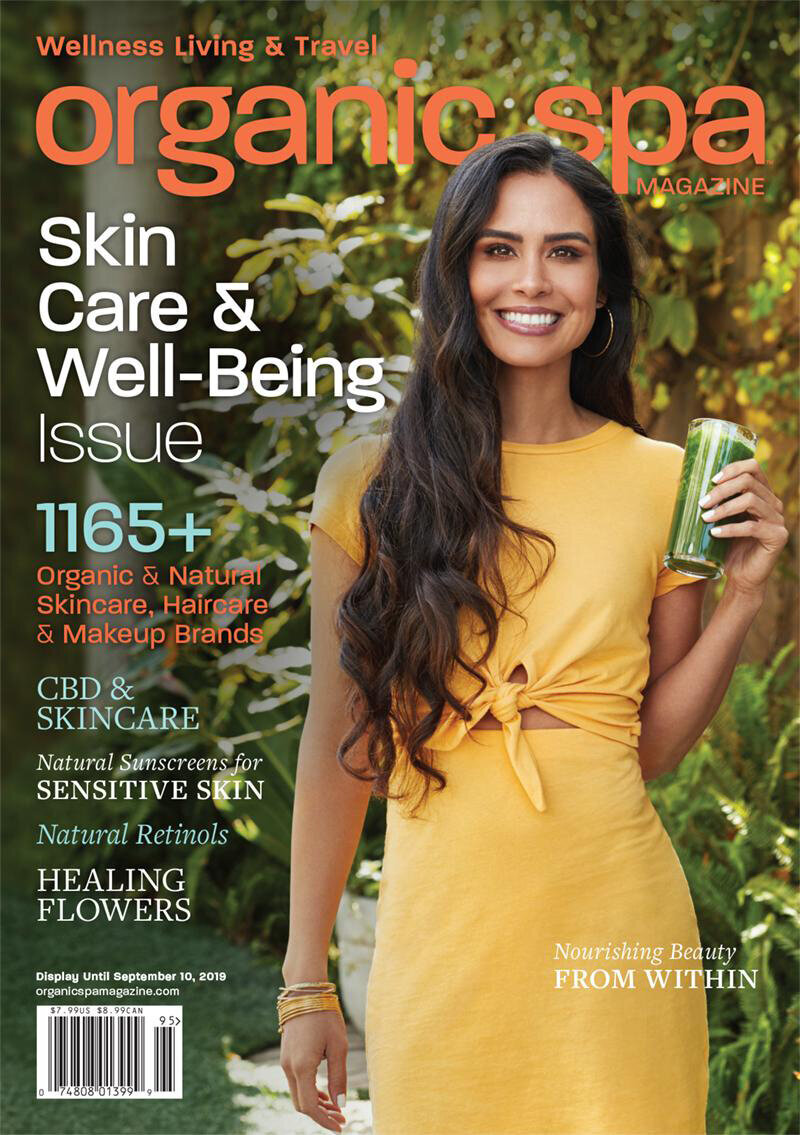 Kimberly Snyder on the cover of Organic Spa Magazine, promoting natural skincare and well-being, holding a green smoothie in a vibrant outdoor setting.