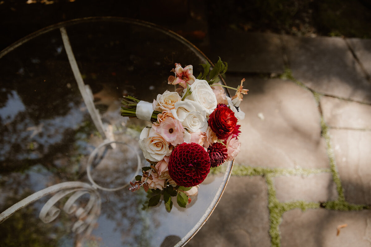A delicate bridal bouquet featuring rich red and soft blush roses, placed elegantly on a glass table reflecting the surrounding garden.