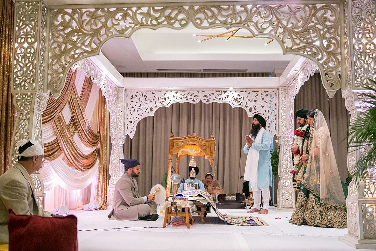 a sheikh groom and hindu bride at an Indian wedding underneath an ivory and gold filigree mandap with a pandit seated in front of swags fo gold drapery