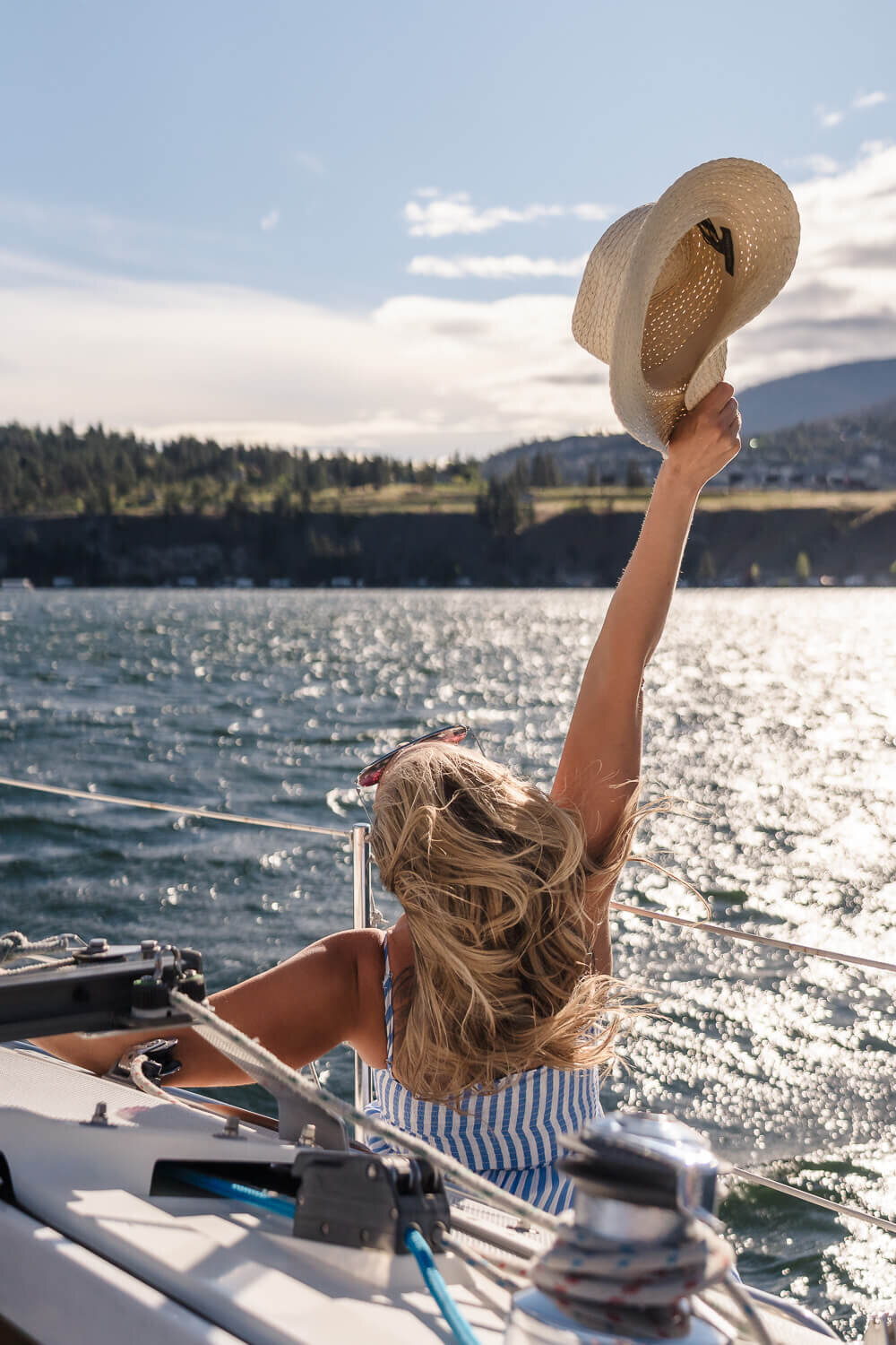 Carefree entrepreneur in striped dress holding a straw hat in the air while sailing on Okanagan Lake.