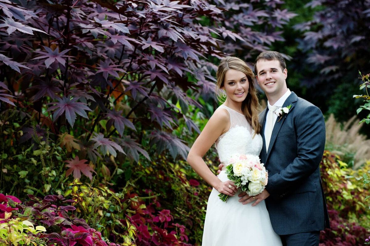 a bride and groom smile at the camera surrounded by colorful foliage in a garden