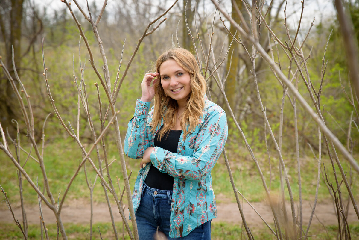 teenage girl is standing in trees with her hand in her hair wearing blue western shirt and jeans at Fonferek Glen in Ledgeview, Wisconsin