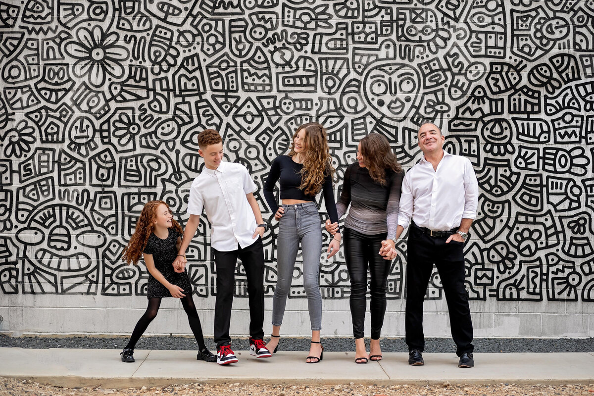 A family of five holds hands and stands in front of a black and white urban mural.